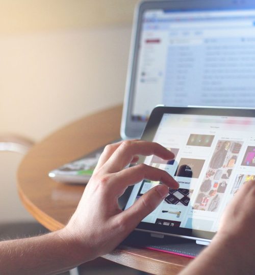 Hands using a tablet beside a laptop in a home office setting, showcasing online browsing.